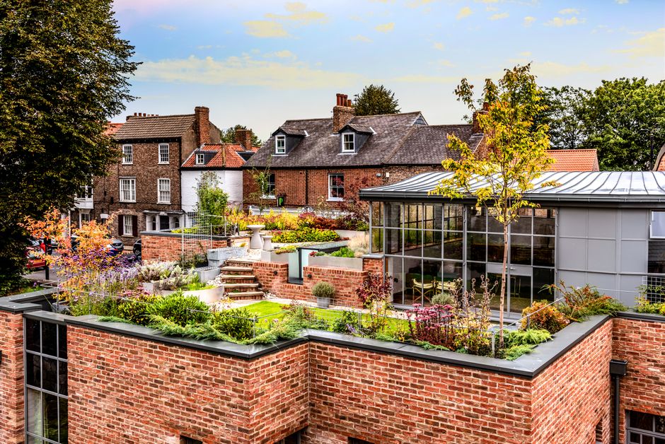 A high-angle view over a dense collection of rooftops in an urban area, featuring various flat and pitched roofs with grey and rust-colored metal sheeting. Among the rooftops, a small rooftop garden i